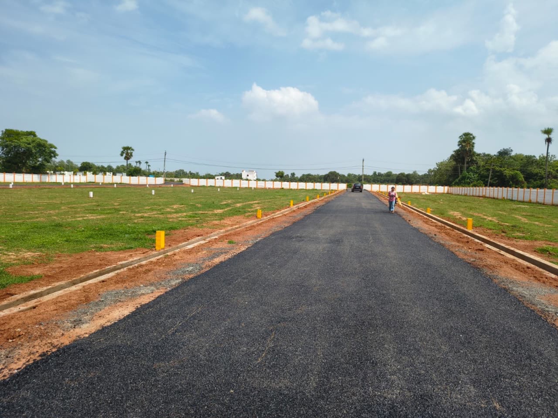 Blacktop internal road running through a developed Kaladhar Infra plotted layout, with demarcated plots and a compound wall on either side